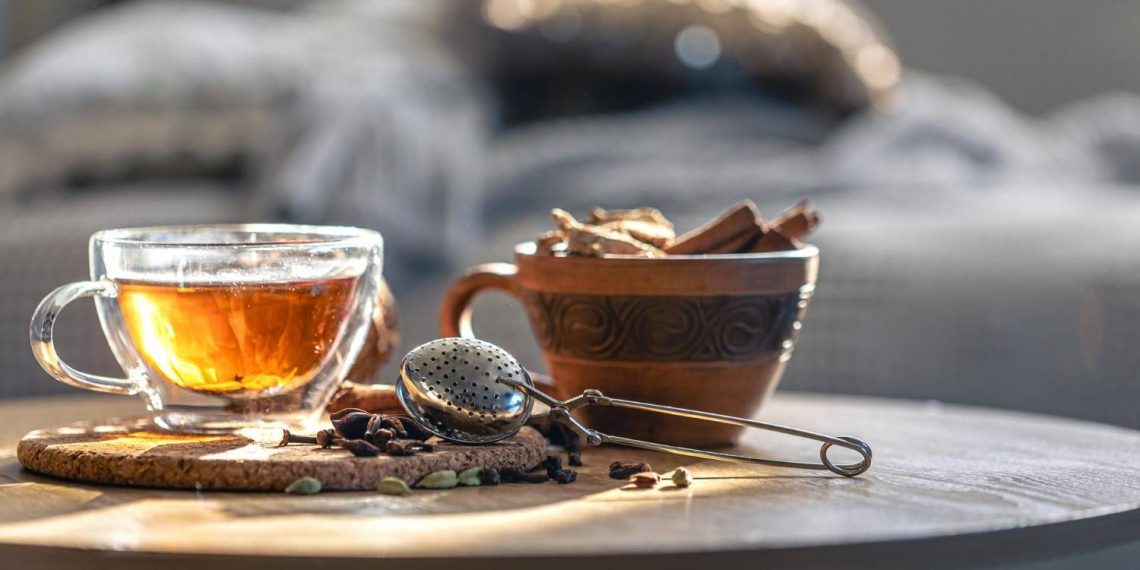 Warm cup of herbal tea with spices and strainer on table.