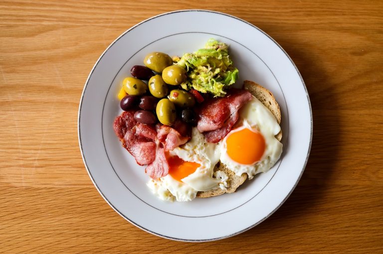 Breakfast plate with fried eggs, bacon, olives, and avocado.