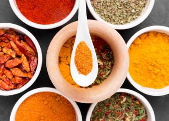 Assorted spices in bowls and a wooden mortar with ground chili on a spoon.