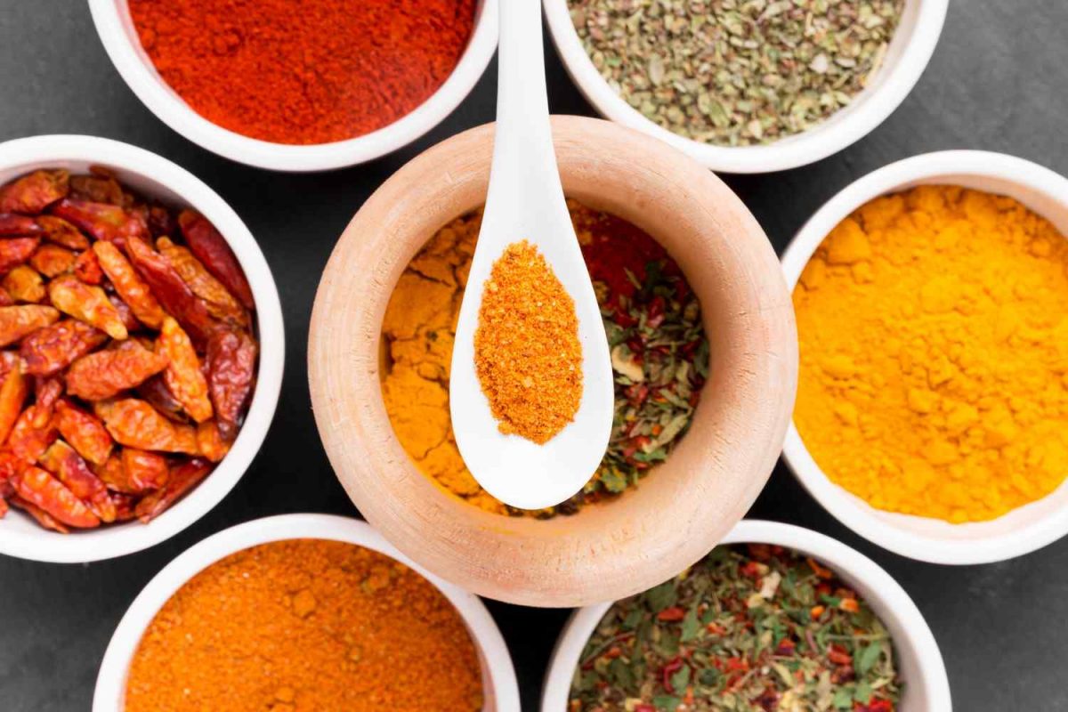 Assorted spices in bowls and a wooden mortar with ground chili on a spoon.