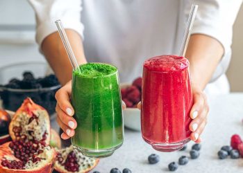 Hands holding green and red smoothies with fresh berries and pomegranates.