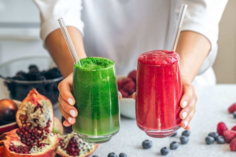 Hands holding green and red smoothies with fresh berries and pomegranates.