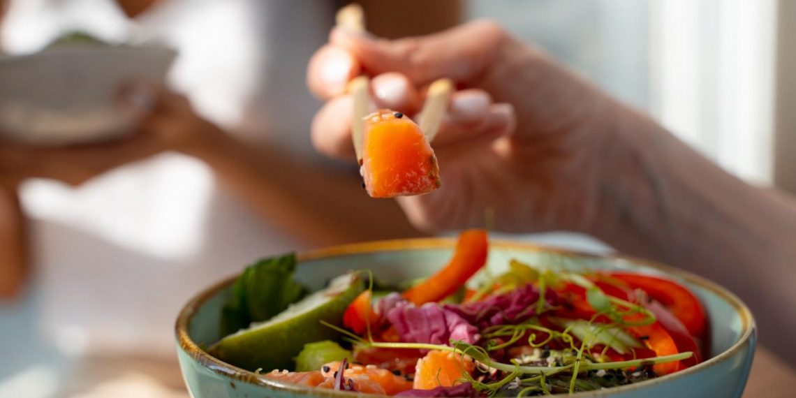 Chopsticks lifting fresh salmon from colorful poke bowl.