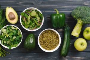 Fresh vegetables and fruits on a wooden table include avocado, greens, and broccoli.