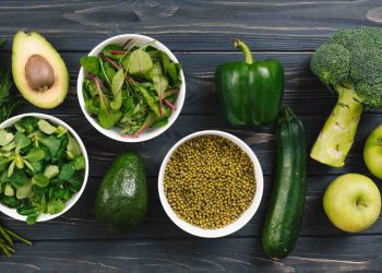 Fresh vegetables and fruits on a wooden table include avocado, greens, and broccoli.