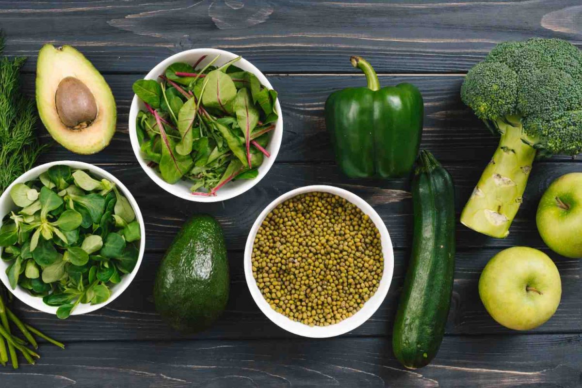 Fresh vegetables and fruits on a wooden table include avocado, greens, and broccoli.