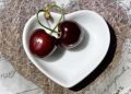 Two cherries in a heart-shaped bowl on a textured surface.