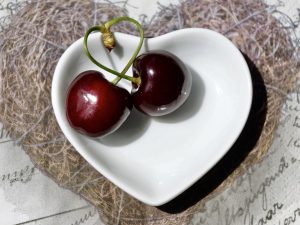 Two cherries in a heart-shaped bowl on a textured surface.