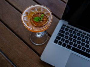 Cocktail garnished with citrus beside a laptop on a wooden table.