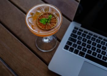 Cocktail garnished with citrus beside a laptop on a wooden table.