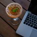 Cocktail garnished with citrus beside a laptop on a wooden table.