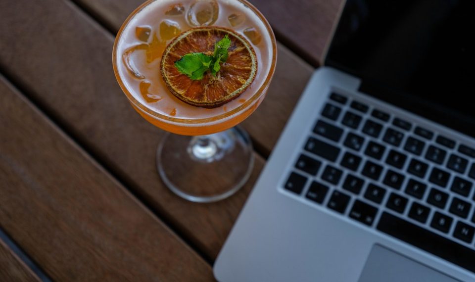 Cocktail garnished with citrus beside a laptop on a wooden table.
