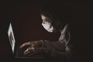 Person wearing mask working intently on a laptop in a dark room.