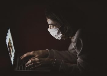 Person wearing mask working intently on a laptop in a dark room.