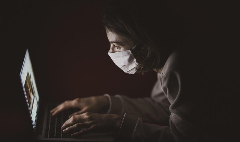 Person wearing mask working intently on a laptop in a dark room.
