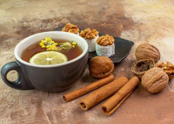 Tea with lemon slice, walnuts, sweets, and cinnamon sticks on textured surface.