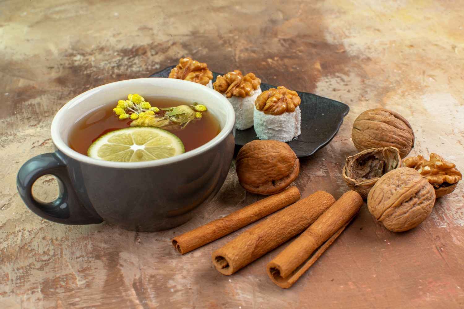 Tea with lemon slice, walnuts, sweets, and cinnamon sticks on textured surface.