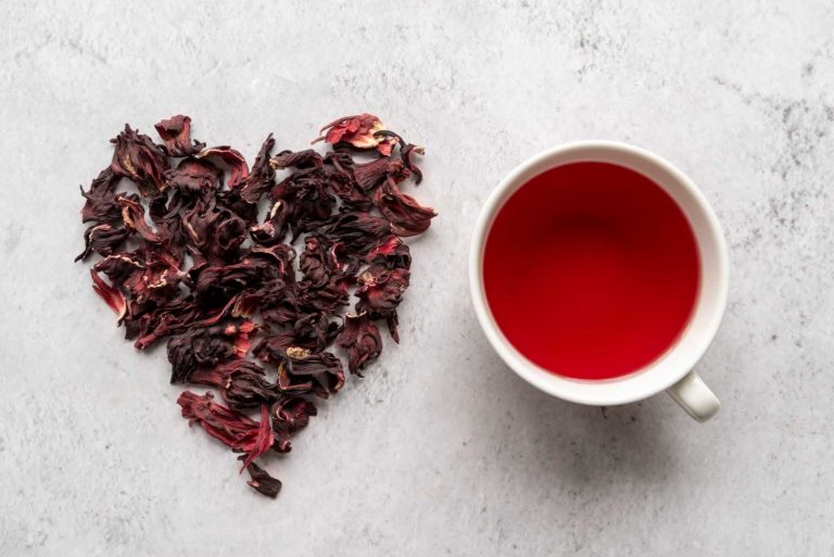 Dried hibiscus petals forming a heart next to a cup of hibiscus tea.