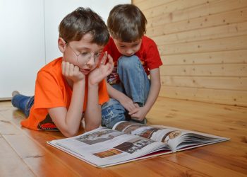 Two boys reading a book on a wooden floor.