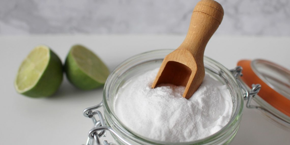 Baking soda in a glass jar with a wooden scoop, next to sliced lime.