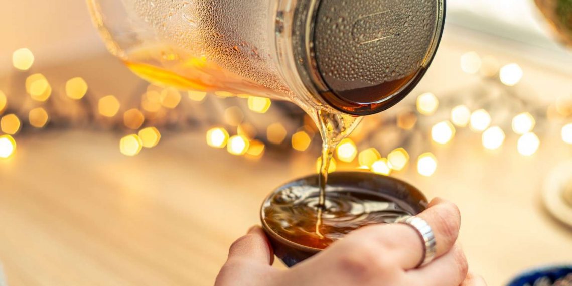 Pouring hot tea from a glass pot into a cup with bokeh lights in the background.