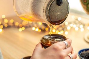 Pouring hot tea from a glass pot into a cup with bokeh lights in the background.
