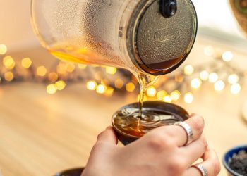 Pouring hot tea from a glass pot into a cup with bokeh lights in the background.