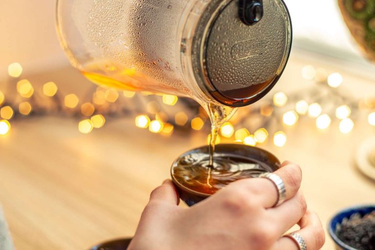 Pouring hot tea from a glass pot into a cup with bokeh lights in the background.