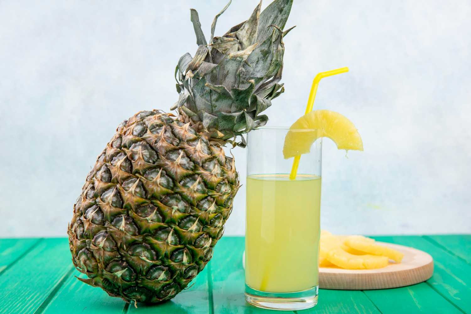 A whole pineapple with its spiky leaves, next to a glass of pineapple juice with a yellow straw and a pineapple slice garnish, and a small wooden board with pineapple slices on a green surface.