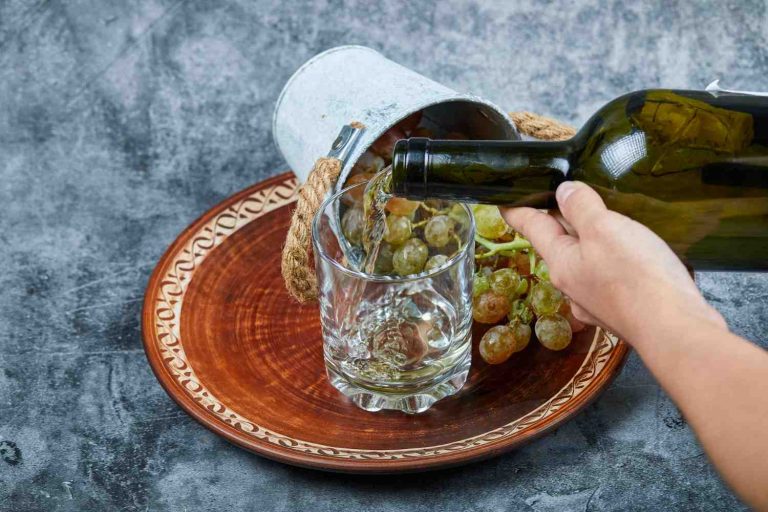 Hand pouring white wine from a green bottle into a glass, with a plate of green grapes and an overturned bucket on a decorative plate.