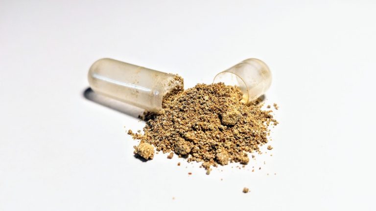 An open, clear capsule spilling granular beige ashwagandha powder onto a white surface.