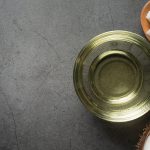 Overhead shot of coconut oil in a glass bowl, with pieces of coconut and a half of a coconut on a grey textured background. The glass bowl is the main focus, with the oil being in a light yellow color. A small wooden bowl with pieces of coconut is in the top right, and the base of a coconut is in the bottom right corner.