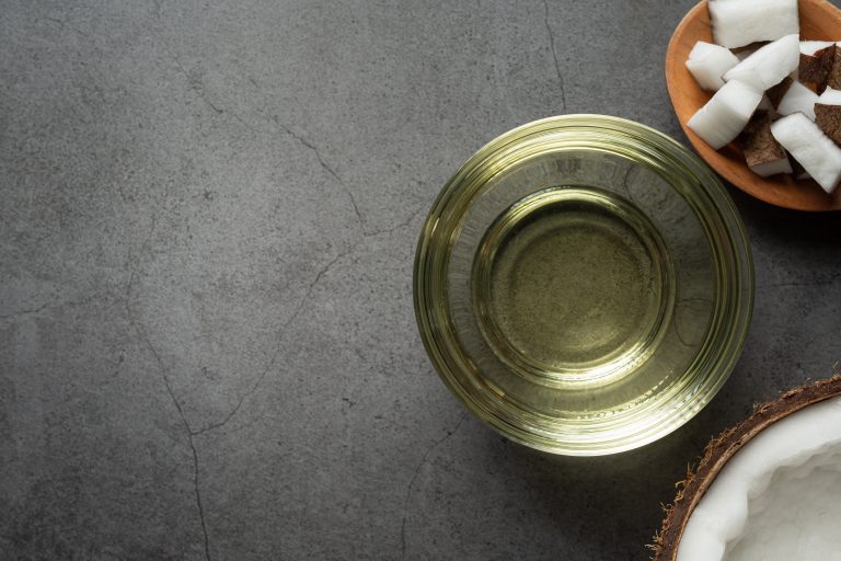 Overhead shot of coconut oil in a glass bowl, with pieces of coconut and a half of a coconut on a grey textured background. The glass bowl is the main focus, with the oil being in a light yellow color. A small wooden bowl with pieces of coconut is in the top right, and the base of a coconut is in the bottom right corner.