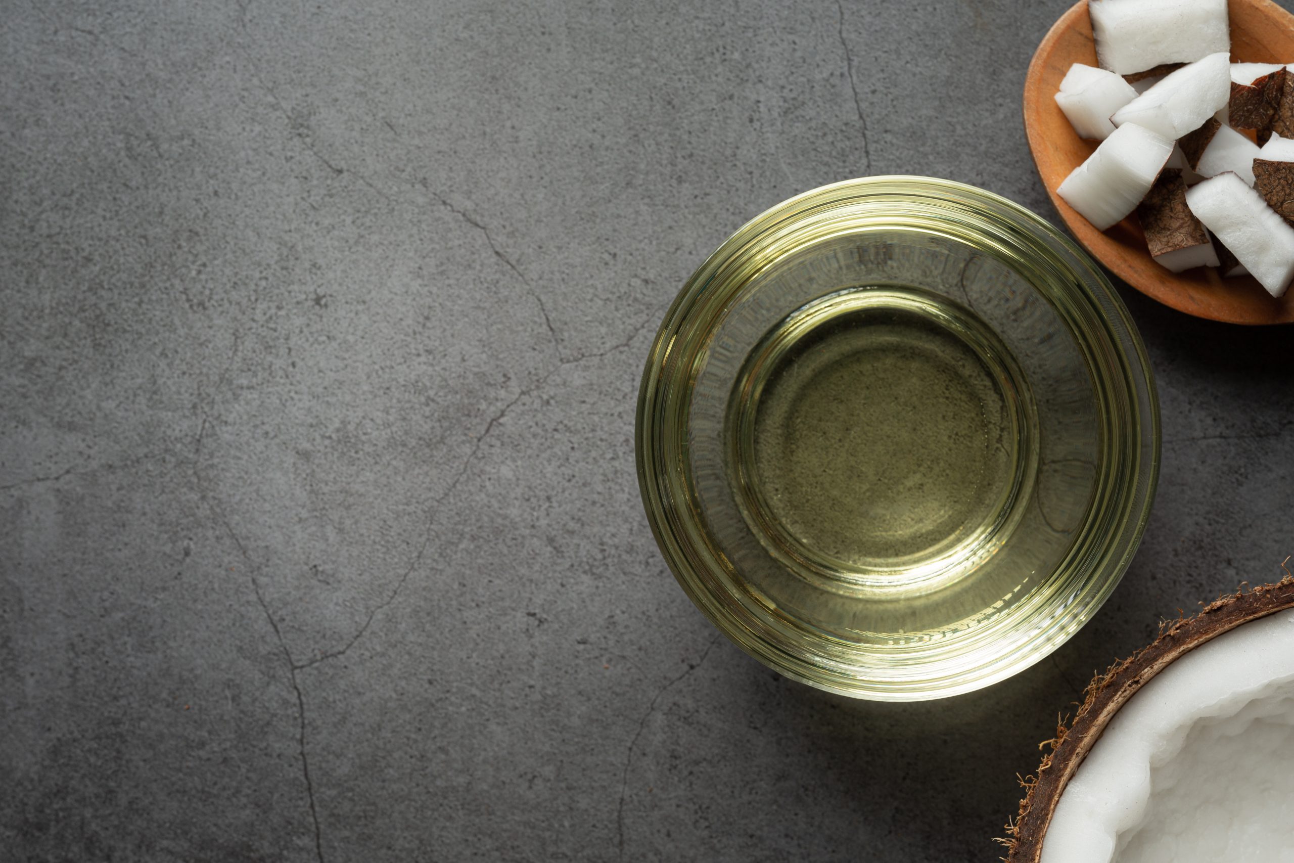 Overhead shot of coconut oil in a glass bowl, with pieces of coconut and a half of a coconut on a grey textured background. The glass bowl is the main focus, with the oil being in a light yellow color. A small wooden bowl with pieces of coconut is in the top right, and the base of a coconut is in the bottom right corner.