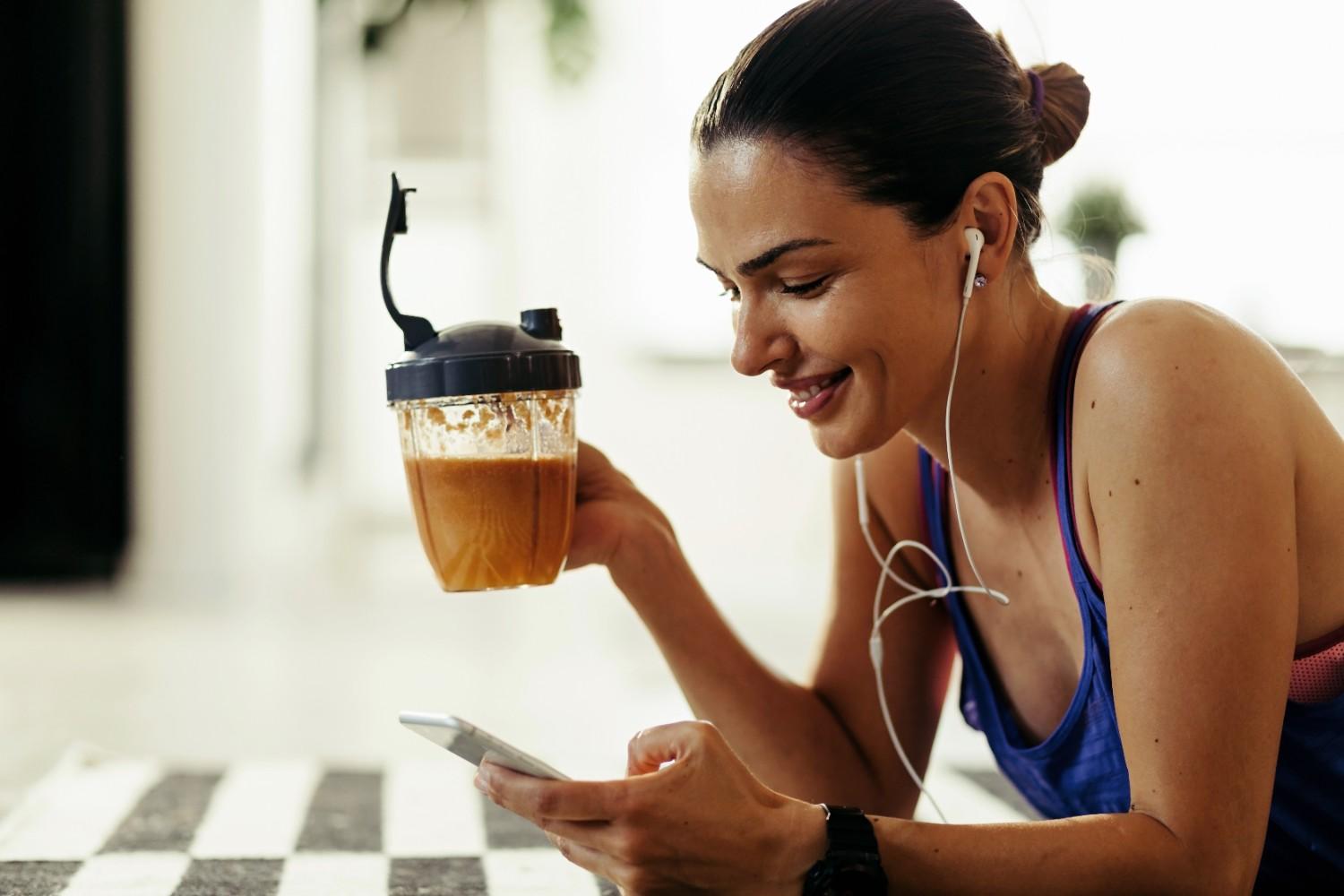 Woman in athletic wear smiling while holding a protein coffee cup, looking at a phone, with earbuds in, on a checkered floor.