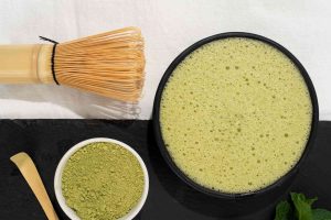 Overhead shot of matcha preparation. A whisk, a bowl of frothy green matcha tea, and a small bowl of green matcha powder with a scoop are arranged on a black slate and a white cloth.