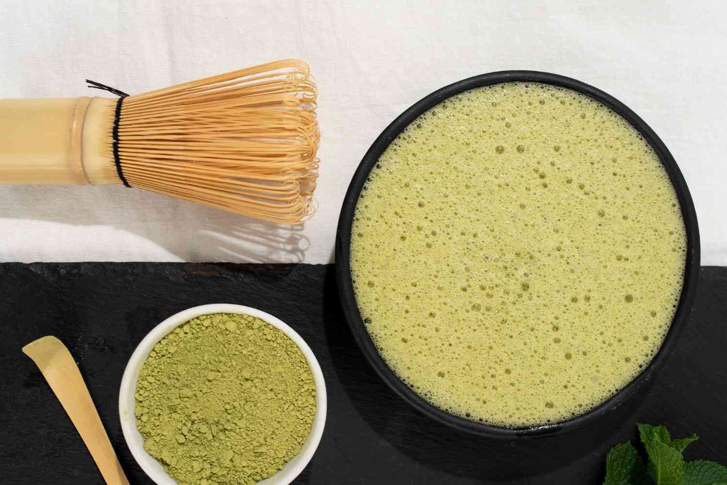 Overhead shot of matcha preparation. A whisk, a bowl of frothy green matcha tea, and a small bowl of green matcha powder with a scoop are arranged on a black slate and a white cloth.