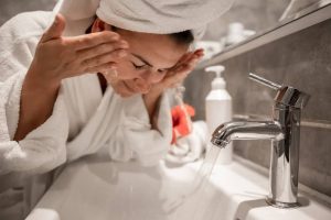 A woman wearing a white robe and a towel on her head washes her face with water flowing from a chrome faucet over a white sink.