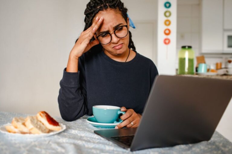 A woman holding her head in frustration while working on a laptop at home, drinking coffee.